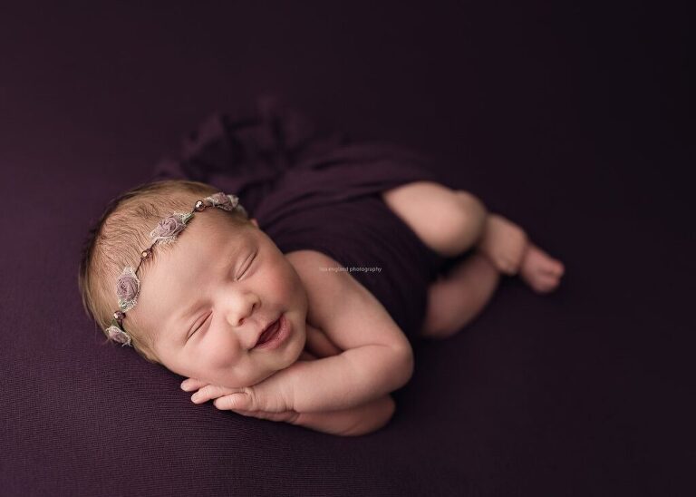 Smiley newborn girl wearing a purple floral headband, sleeping on her side on a purple blanket - Lisa England Photography, San Diego Newborn Photography