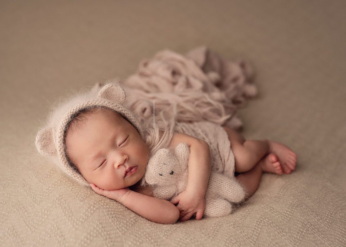 newborn baby girl wearing a fuzzy, blush pink teddy bear bonnet, sleeping on her side holding a tiny matching teddy bear - Lisa England Photography, San Diego baby photographer