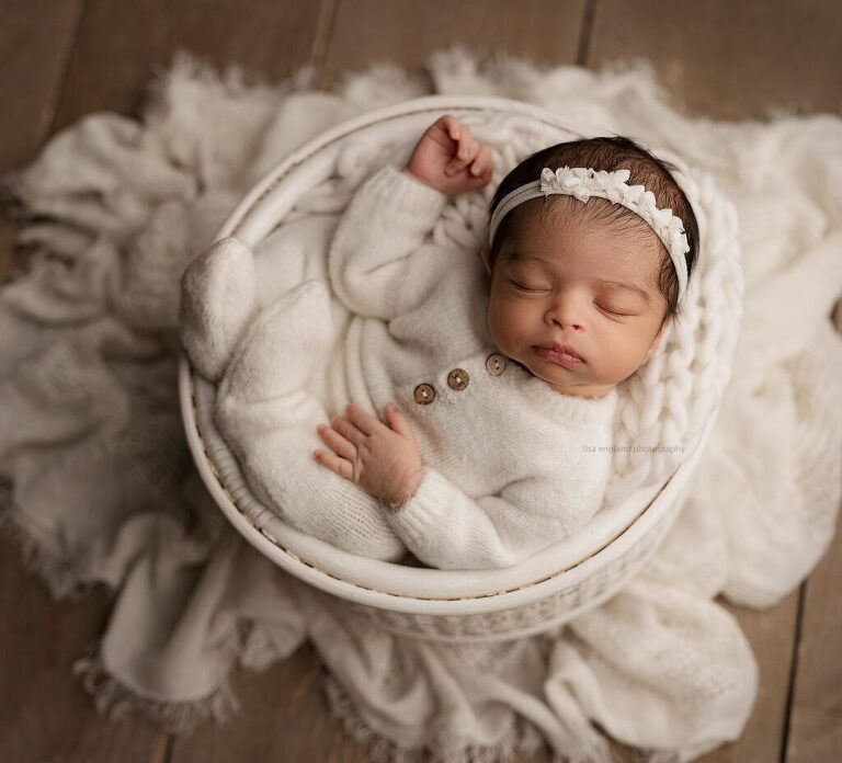newborn baby girl wearing a white outfit with a white bows tieback, sleeping soundly in a white round basket - Lisa England Photography, studio newborn photographer in San Diego